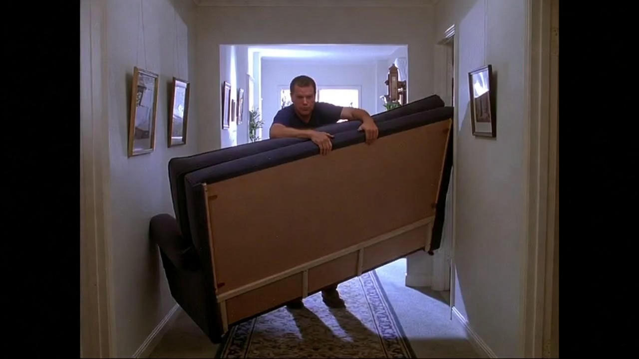 Comedic scene of a tall character struggling to move a couch in a narrow hallway, referencing Ross’s famous Pivot moment.